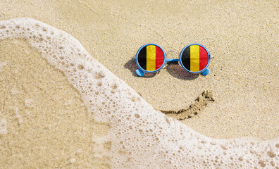 Fototapeta na wymiar Sunglasses with flag of the Belgium on a sandy beach. Nearby is a sea lightning and a painted smile.