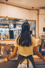 Young female barista in apron standing behind the counter bar of coffee shop. Waitress bartender...