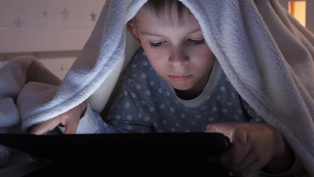 Portrait of boy in pajamas lying under blanket and using tablet computer. Children education, development, kids using gadgets secrecy, privacy.