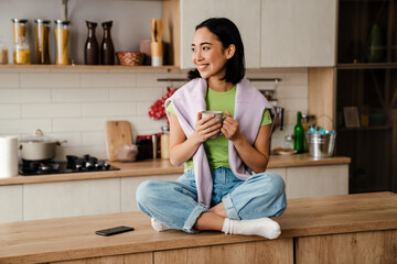 Cheerful young asian woman drinking coffee while sitting in cozy kitchen
