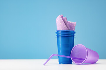 Plastic blue glasses with paper napkins, lilac glass with a drinking straw, blue background. Plastic recycling concept.