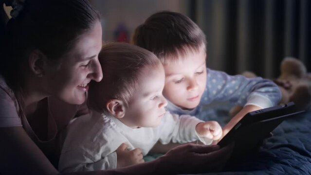 Two cute boys in pajamas using tablet computer in bed with mother. Family having time together, parenting, happy childhood and entertainment