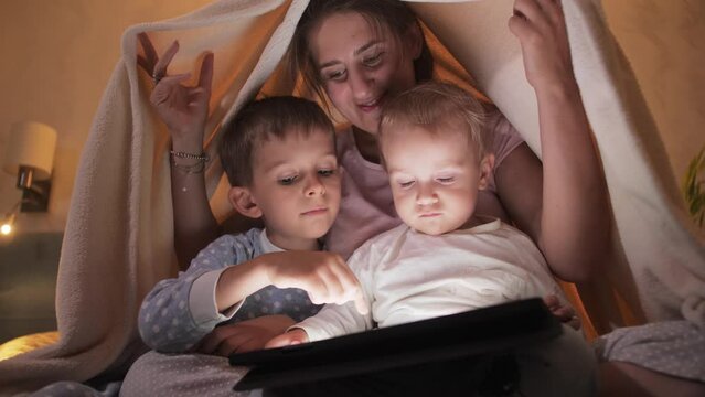 Happy smiling mother with two sons in pajamas having fun on bed and playing games on tablet computer. Family having time together, parenting, happy childhood and entertainment
