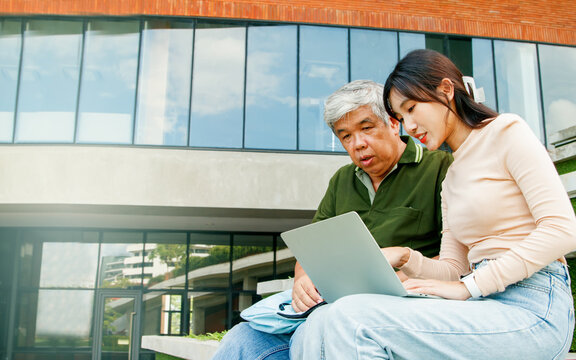 Daughter and father sit together in the vacant space on campus spend their free time teaching senior father to learn how to read health care for old age father understand simple use of social media.