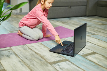 Little girl watching online video on laptop and doing stretching, fitness exercises at home