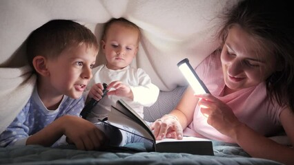 Young loving mother reading bedtime story book to her two sons hiding under blanket in bed. Family having time together, parenting, happy childhood and entertainment