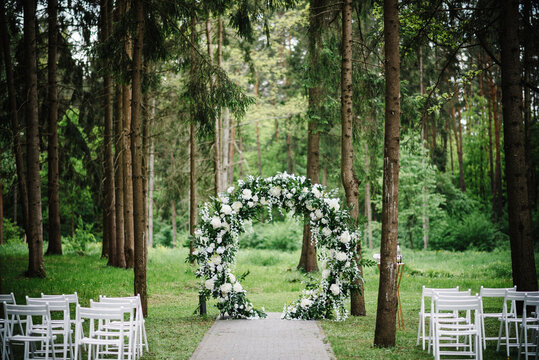 Wedding ceremony in the forest. The road to the round arch with foliage, greens, greenery, and flowers. Rustic decor. Wooden chairs in the backyard banquet area. Seats for guests.