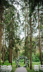 Wedding ceremony in the forest. The road to the round arch with foliage, greens, greenery, and flowers. Rustic decor. Wooden chairs in the backyard banquet area. Seats for guests.