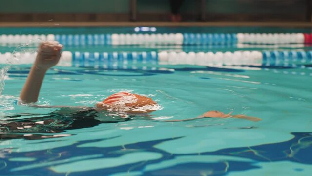 Beautiful Little Girl Trains To Swim Front Crawl With Aquatic Dumbbell. Child To Learn To Swim Changing Hands In The Swimming Pool. Low Angle Side View Tracking Shot