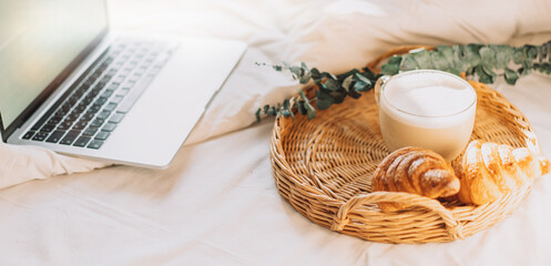 Fototapeta na wymiar Wicker tray with croissants and coffee on white bed linen with laptop.