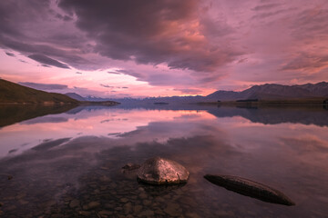 Fototapeta premium Sunrise over a mirror calm Lake Tekapo and snow capped mountains in New Zealand with rocks in the foreground