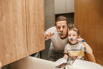 Reflection of the little boy and his father brushing their teeth together in a bathroom.