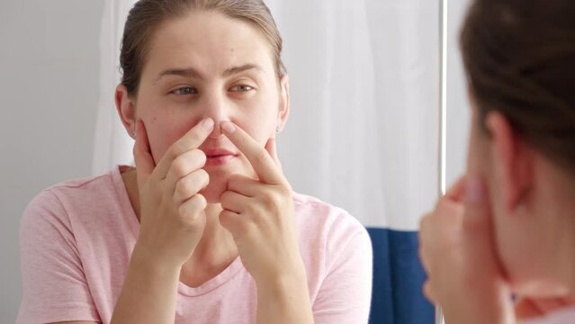 Young woman looking in mirror and squeezing pimples on her nose. Concept of female aging, skin issues, facial treatment and care