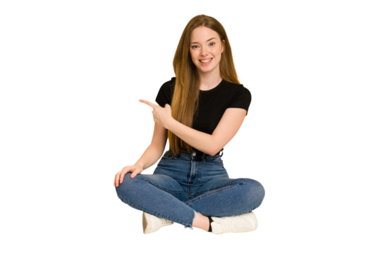 Young redhead woman sitting on the floor cut out isolated smiling and pointing aside, showing something at blank space.