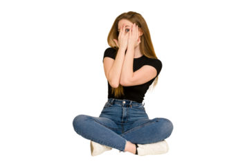 Young redhead woman sitting on the floor cut out isolated blink through fingers frightened and nervous.