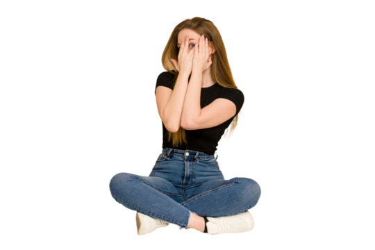 Young redhead woman sitting on the floor cut out isolated blink through fingers frightened and nervous.