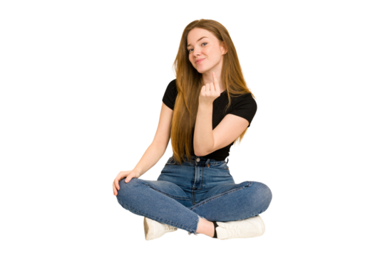 Young redhead woman sitting on the floor cut out isolated pointing with finger at you as if inviting come closer.