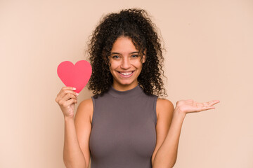 Young african american woman holding a heart for valentines day isolated showing a copy space on a palm and holding another hand on waist.