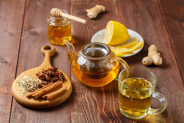 Obraz na płótnie Canvas A transparent mug of lemongrass tea on a table next to anise, cinnamon, ginger and honey and a teapot with tea on a wooden board.