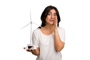 Young Indian woman holding a small wind energy mill isolated is saying a secret hot braking news...