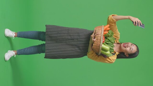 Full Body Of Asian Female Farmer With Vegetable Basket Taking Selfie By Smart Phone On Green Screen Background In The Studio
