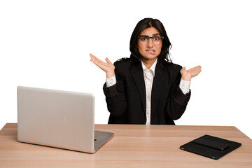 Young indian woman in a table with a laptop and tablet isolated confused and doubtful shrugging shoulders to hold a copy space.