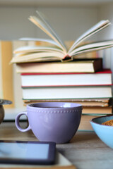 Stack of books, e-reader, reading glasses, bowl of biscuits, apples and cup of tea on the table. Bookshelf in the background. Selective focus.