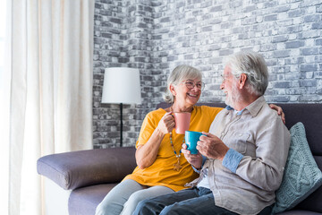 Happy mature 50s husband and wife sit rest on comfortable sofa in living room enjoy tea talking,...