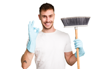 Young man holding a broom to clean his house cut out isolated cheerful and confident showing ok gesture.