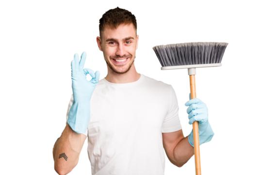 Young man holding a broom to clean his house cut out isolated cheerful and confident showing ok gesture.