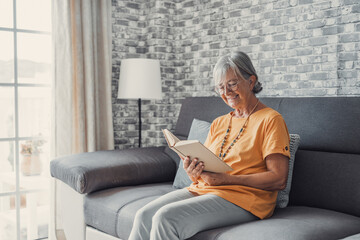Obraz premium Happy older blonde woman sitting on comfortable sofa in living room, reading interesting book. Smiling elder retired grandmother enjoying leisure weekend time with favorite novel alone at home..
