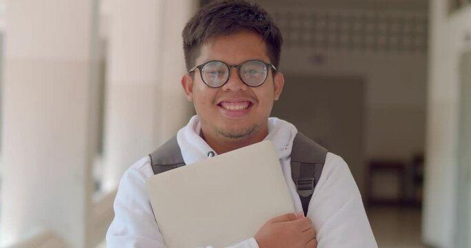 A happy smile of an Asian male teenage high school student wearing thick glasses. wearing a white sweater, carrying a laptop with a backpack standing on the corridor in the building.