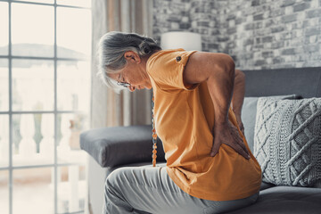 Unhappy mature woman touching back, feeling pain, sitting on couch in living room, unhealthy middle...
