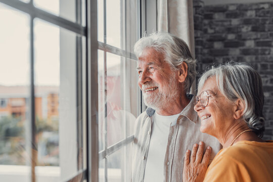 Happy Bonding Loving Middle Aged Senior Retired Couple Standing Near Window, Looking In Distance, Recollecting Good Memories Or Planning Common Future, Enjoying Peaceful Moment Together At Home..