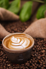 Cappuccino in a ceramic cup with flower or plant drawn on the foam and roasted coffee beans or seeds scattered on the table and in a jute sack, with green plant in background