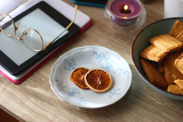 Notebook, pen, tablet, reading glasses, books, phone, candle, bowl of biscuits and cup of tea on the table. Selective focus.