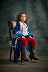 Portrait of little charming girl with curly hair in image of little prince posing over on dark green background. Concept of historical remake, comparison of eras, retro, vintage, emotions