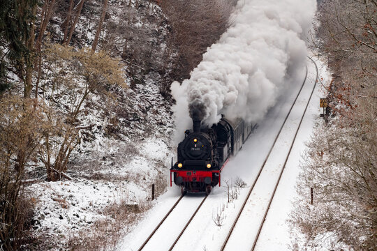 Steam train on the “Ruhrtalbahn“ (Ruhr valley line) between Arnsberg and Meschede Sauerland Germany heading to Winterberg. Engine with big puff of smoke on snowy winters day in idyllic landscape.