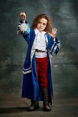 Fototapeta premium Surprised little girl with curly hair in image of little prince holding toy spider over dark green background. Concept of historical remake, comparison of eras, retro, vintage, emotions