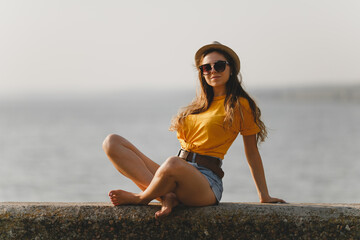Portrait of young sexy girl in hat, yellow bright t-shirt, denim shorts, sunglasses sitting at...