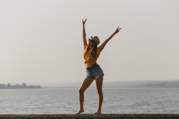Fototapeta na wymiar Young sensual woman in hat, bright yellow t-shirt, denim shorts, sunglasses standing at lake parapet smiling showing victory symbol with hands 