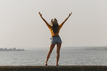 Young sexy woman in yellow t-shirt, denim shorts, hat is standing at lake beach with raised up hands; freedom concept