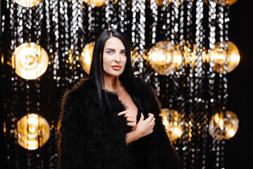 Young beautiful smiling brunette female in trendy evening fur coat. Carefree woman posing near shiny tinsel wall in studio. Fashionable model with bright makeup looks at camera