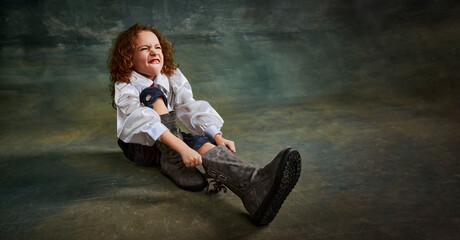 Wide angle view of little cute kid with curly hair sitting on floor over dark green background. Concept of childhood, comparison of eras, retro, vintage, emotions