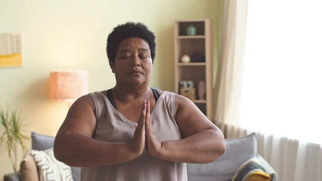African American mature woman making hands in prayer position while meditating in calm domestic atmosphere at daytime