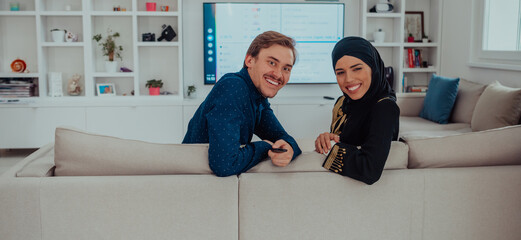 Young muslim couple woman wearing islamic hijab clothes sitting on sofa watching TV together during the month of Ramadan at modern home
