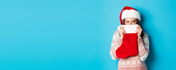 Cute girl cover face with christmas stocking, staring right with cunning gaze, standing in Santa...