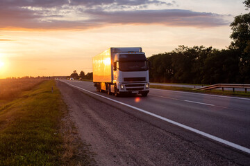 Obraz na płótnie Canvas A white truck with a semitrailer transports cargo on the highway in the evening against the backdrop of sunset. Compliance with the rules for the carriage of goods. Transport inspection, european