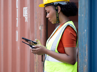 Female foreman worker wears yellow safety hardhat holding radio and mobile phone to control loading cargo box at container yard, portrait. Multiracial engineer woman works at international terminal