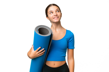 Teenager caucasian girl going to yoga classes while holding a mat over isolated background looking up while smiling